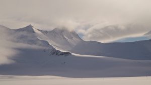 Snow-covered mountains partially shrouded in mist and clouds under a softly lit sky, with a vast, flat expanse of snow in the foreground.