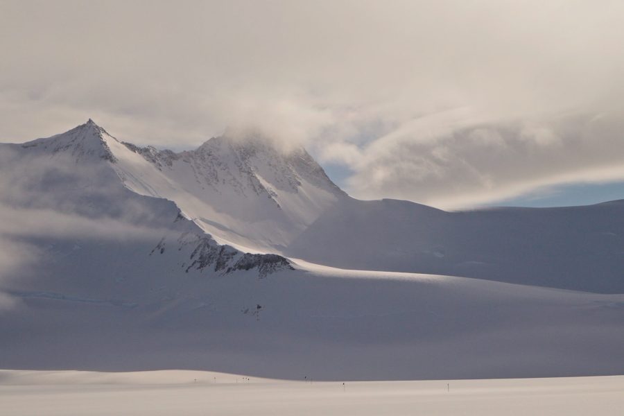 Snow-covered mountains partially shrouded in mist and clouds under a softly lit sky, with a vast, flat expanse of snow in the foreground.