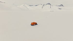 An orange vehicle travels across a vast, snowy landscape with distant snow-covered mountains under an overcast sky. The scene appears remote and isolated.