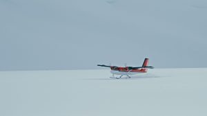 A small red and white propeller plane is landing or taking off on a vast, snowy landscape under an overcast sky.