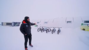 A person in winter clothing stands in snowy conditions, gesturing toward several bicycles lined up outside a row of white, tent-like structures. Snow covers the ground and the surroundings.