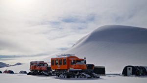 Two orange snow vehicles with treads are parked on a snowy landscape near a small black shelter, with a large snow-covered hill in the background under a cloudy sky.