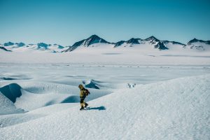 A person in winter gear walks alone across a vast, snow-covered Antarctic landscape, with distant jagged mountains under a clear blue sky—echoing the spirit of Antarctic Logistics and Expeditions.
