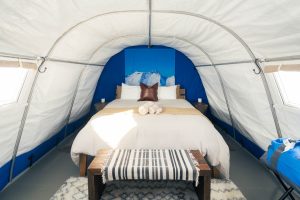 A cozy glamping tent interior with a neatly made queen bed, white and beige linens, folded towels, a brown pillow, a bench with a striped blanket, and blue walls featuring a mountain print behind the bed.