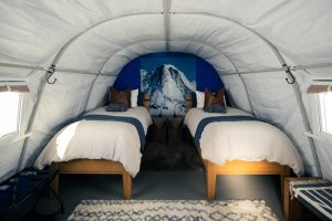Two single beds with white bedding and brown pillows are inside a white, arched tent. A large photo of a snow-covered mountain hangs on the back wall between the beds. Natural light comes from side windows.
