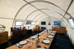 A long dining table set for a meal is inside a large, white, insulated tent with curved ceilings. Cabinets, drinks, and a clock are visible, and windows let in natural light. The space looks cozy and modern.