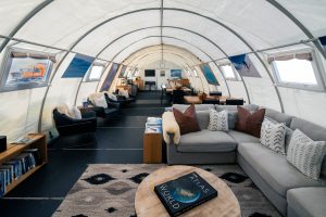 A cozy, modern lounge inside a large, white tent features a gray sectional sofa, armchairs, bookshelves, and pictures on the walls. A round table with an atlas book sits on a patterned rug in the foreground.