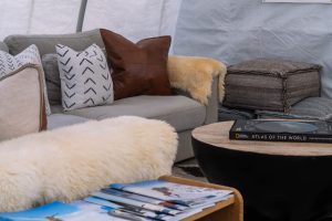 A cozy living room with a gray sofa, patterned and brown pillows, a fur throw, stacked gray cushions, a round wooden coffee table with a book, and a side table with magazines.