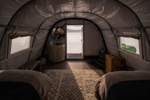 Interior of a tent with two beds, a soft rug, and a small vanity, looking out through an open door to a snowy landscape and cloudy sky. Soft natural light fills the cozy space.