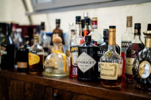Various bottles of liquor, including gin, whiskey, and other spirits, are arranged on a wooden surface in a bar or home setting. The bottles are different shapes, sizes, and brands.
