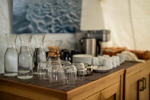 A wooden countertop displays glass carafes, upside-down glasses, stacked plates, cups, and a coffee maker, with baskets and a coffee pot, set against a blurred wall with artwork.