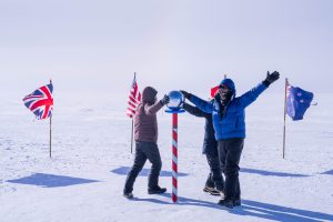 Three people in winter gear stand in a snowy landscape around a striped pole topped with a shiny sphere, with British, American, and New Zealand flags in the background.