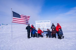 Nine people in winter gear pose next to a large Geographic South Pole sign and a U.S. flag, standing on snow under a blue sky in Antarctica.