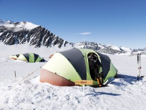 A person in cold-weather gear stands at the entrance of a green tent labeled Byrd in Antarctica, surrounded by snow and mountains under a clear blue sky. Other tents and Antarctic Logistics and Expeditions equipment dot the wintry landscape.
