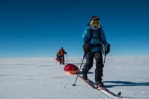 Four people dressed in cold-weather gear ski across a snowy, flat landscape under a clear blue sky in Antarctica, pulling sleds with their equipment behind them as part of an Antarctic Logistics and Expeditions journey.