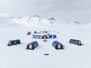 Aerial view of a remote snow-covered camp with several blue and black cabins, parked vehicles, and mountains in the background under an overcast sky.