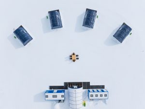 Aerial view of five dark-roofed tents arranged in a semicircle on snow around a central table with chairs, near two rectangular white buildings at the bottom of the image.