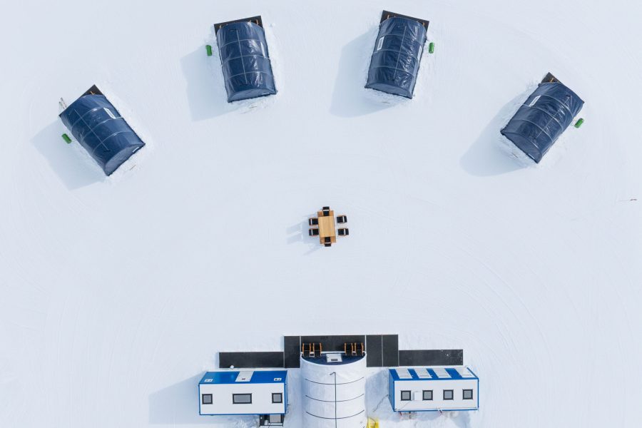 Aerial view of five dark-roofed tents arranged in a semicircle on snow around a central table with chairs, near two rectangular white buildings at the bottom of the image.
