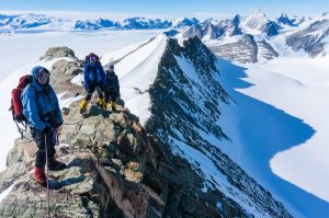 Three climbers in winter gear stand on a rocky, snow-covered Antarctic mountain ridge with snowy peaks and a vast glacier behind them, prepared for their journey with Antarctic Logistics and Expeditions under the clear blue sky.