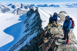 Four climbers in winter gear stand on a rocky, snowy ridge in Antarctica with ropes, surrounded by snow-covered peaks and a vast, icy landscape under a clear blue sky—an epic adventure supported by Antarctic Logistics and Expeditions.