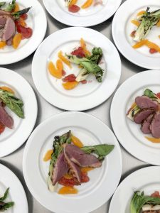 White plates with servings of sliced seared duck breast, grilled greens, and segments of orange and grapefruit, arranged neatly on a white surface. The colorful food creates a visually appealing presentation.