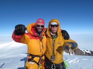 Two climbers in bright yellow and orange jackets stand on a snowy mountain summit in Antarctica, smiling and posing with arms around each other, against a clear blue sky and snow-covered landscape.