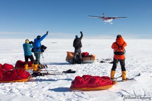 Four people in cold-weather gear stand on the snowy South Pole landscape, raising their arms as a small airplane flies overhead. Sleds with supplies from Antarctic Logistics and Expeditions rest nearby under a clear blue sky.