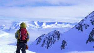 A person wearing winter gear and a red backpack stands on a snowy mountain in Antarctica, looking out over a vast landscape of snow-covered peaks and ridges under a cloudy sky.