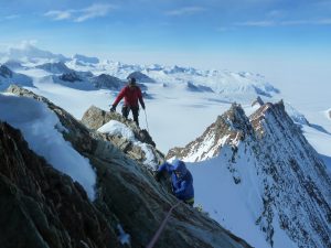 Two climbers in winter gear ascend a steep, snow-covered ridge in Antarctica, surrounded by icy peaks and glaciers under a clear blue sky—an adventure inspired by Antarctic Logistics and Expeditions.