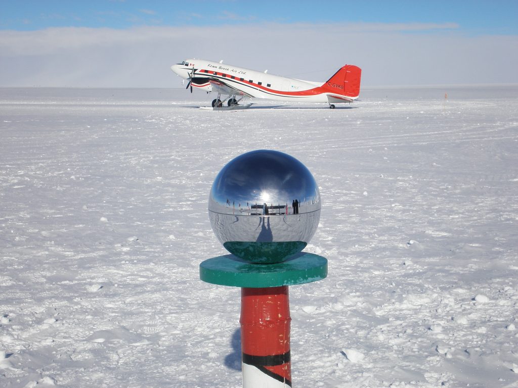 A shiny silver sphere on a pole stands in the snowy South Pole landscape, reflecting people and surroundings; in the background, a red and white propeller plane sits on the snow under a brilliant Antarctic sky.