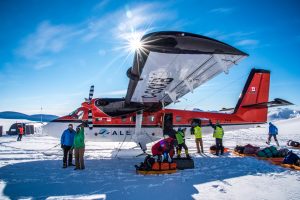 People unload equipment from a red and white plane on the snowy expanse of Antarctica under a clear blue sky. The sun shines brightly as several individuals in colorful jackets and safety vests work with Antarctic Logistics and Expeditions.