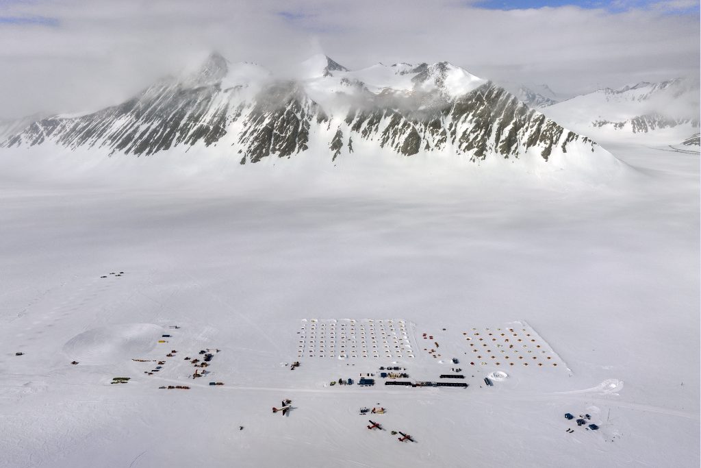 Aerial view of a snowy mountain range in Antarctica with a large camp below, featuring rows of orange tents and scattered equipment on a vast, icy plain. This South Pole site is supported by Antarctic Logistics and Expeditions under a partly cloudy sky.