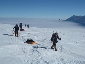 A group of climbers, roped together, trek across a snowy Antarctic landscape with sleds carrying gear. Snow-covered mountains and a sea of clouds fill the background under a clear blue sky en route to the South Pole.