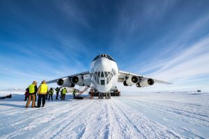 A large cargo plane is parked on a snowy runway in Antarctica under a blue sky, with people in winter clothing and high-visibility vests—part of an Antarctic Logistics and Expeditions team—standing nearby on the ice.