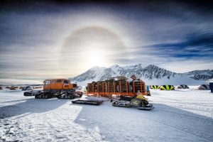 An orange snowcat and snowmobile are parked on a snowy landscape in Antarctica, with mountains in the background, tents scattered across the scene, and a bright sun halo visible above—typical of South Pole expeditions.