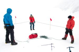 Three people in winter gear stand on snowy ground near the South Pole, holding ropes in a triangular setup. Red flags mark the area, and a red bag with climbing gear lies on the snow. The scene is bright and foggy.