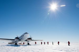 A group of people in red jackets stand on snowy ground near a propeller airplane under a bright, sunny sky with lens flare, preparing for an Antarctic Logistics and Expeditions journey in Antarctica.