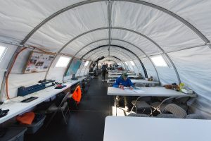 Interior of a large, white, arched tent at the South Pole with long tables and chairs. One person reads at a table in the foreground; equipment, maps, and Antarctic Logistics and Expeditions supplies line the walls. More people are further inside.