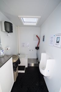 A clean, compact restroom at the South Pole with a toilet, urinal, waste bin, hand sanitizer, and wall-mounted instructions. A skylight brightens the space, while a red hose sits beside a small sink—ideal for Antarctic Logistics and Expeditions.