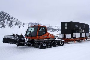 An orange snowcat vehicle with tracks, used by Antarctic Logistics and Expeditions, pulls a black mobile building on a sled across the snowy landscape of Antarctica, with mountains visible in the background under an overcast sky.