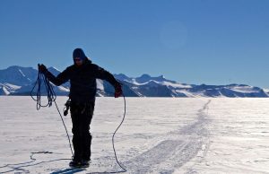 A person dressed in winter gear stands on a snowy, flat landscape in Antarctica with mountains in the distance, coiling a blue rope; a long track stretches behind them in the snow under a clear South Pole sky during an Antarctic Logistics and Expeditions journey.