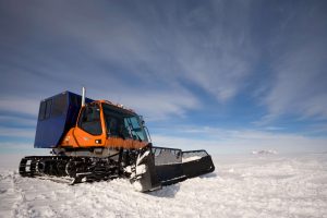 An orange and blue snowcat vehicle with tracks and a front plow sits on a snowy Antarctic landscape, ready for Antarctic Logistics and Expeditions under a partly cloudy sky.