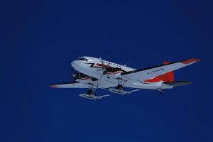 A white and red airplane with skis attached to its landing gear flies against a clear deep blue sky over Antarctica. The aircraft, supporting Antarctic Logistics and Expeditions, has C-GVKB and Kenn Borek Air Ltd. written on its side.