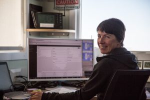 A person with short dark hair sits at a desk, smiling at the camera, with a large computer monitor displaying data sheets—perhaps planning Antarctic Logistics and Expeditions. A Caution: Otter Crossing sign is visible on the wall behind them.