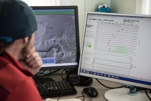 A person looks at two computer monitors; one displays a grayscale map, possibly of Antarctica or the South Pole, while the other shows complex weather charts. A small Cookie Monster toy perches atop the right monitor.