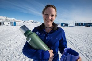 A woman in a blue jacket stands outside in Antarctica, smiling and holding a green thermos and a blue mug. Snow-covered mountains, portable buildings, and the clear sky hint at an Antarctic Logistics and Expeditions site.