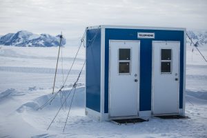 A small blue and white booth with two doors labeled TELEPHONE stands on a snowy landscape at the South Pole, with snow-covered mountains in the background, serving as a unique landmark for Antarctic Logistics and Expeditions in Antarctica.