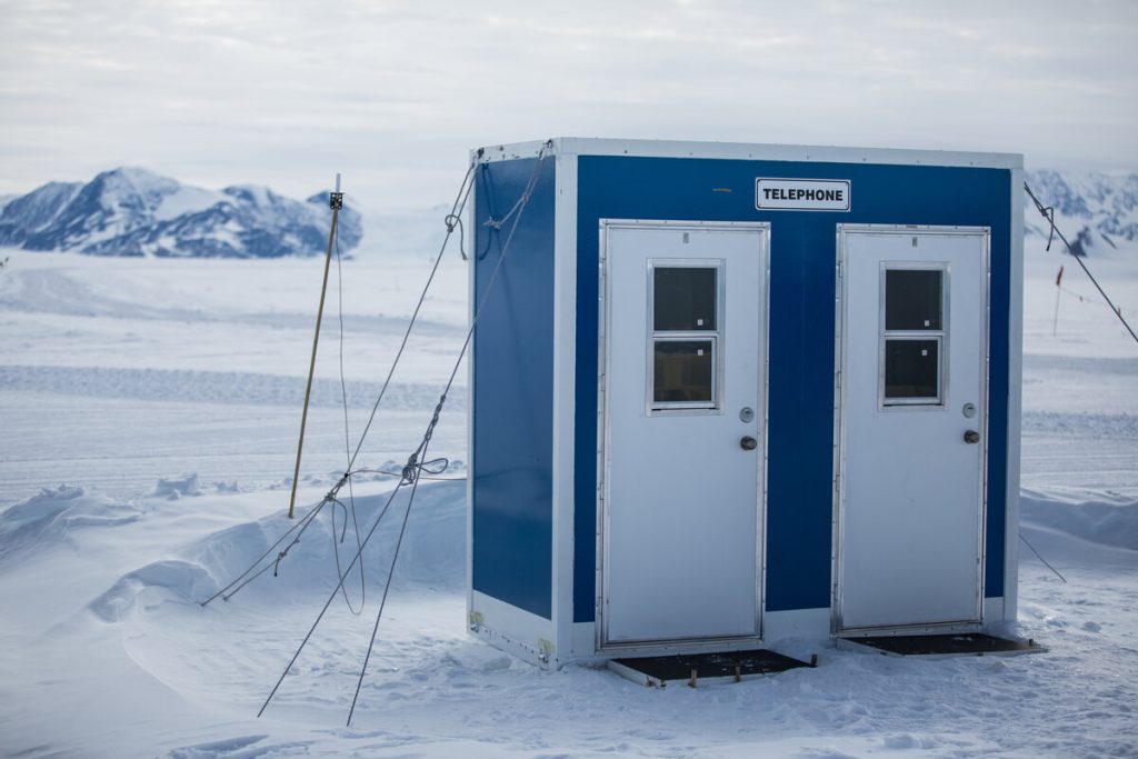A blue and white structure labeled TELEPHONE with two doors stands on a snowy Antarctic landscape near the South Pole, surrounded by mountains and covered in snow, with cables anchoring it to the ground.