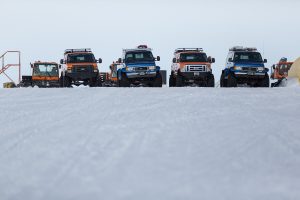 Five off-road vehicles, including trucks and utility vehicles, are parked on a snowy, icy surface in Antarctica under a cloudy sky. Equipped for rugged, cold-weather conditions, they support Antarctic Logistics and Expeditions at the South Pole.