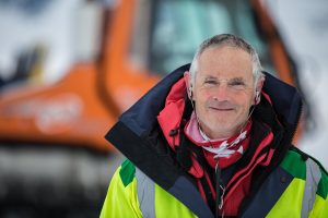 A man in a bright yellow and red jacket stands outdoors in snowy Antarctica, smiling. He wears earphones and a red neck gaiter. An orange snow vehicle, used by Antarctic Logistics and Expeditions, is blurred in the background.